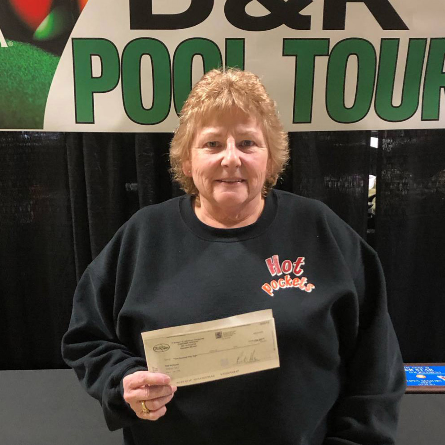 Woman in black sweatshirt holding a check in front of a banner that reads 
