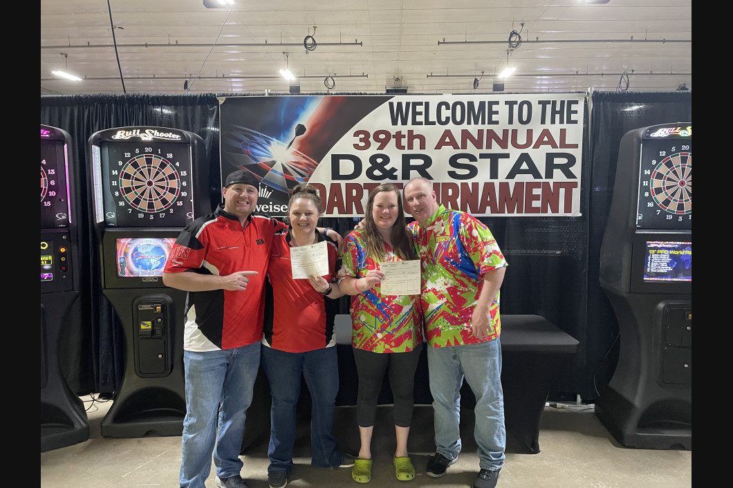 Four people holding certificates at a dart tournament. Black backdrop, dart machines. 