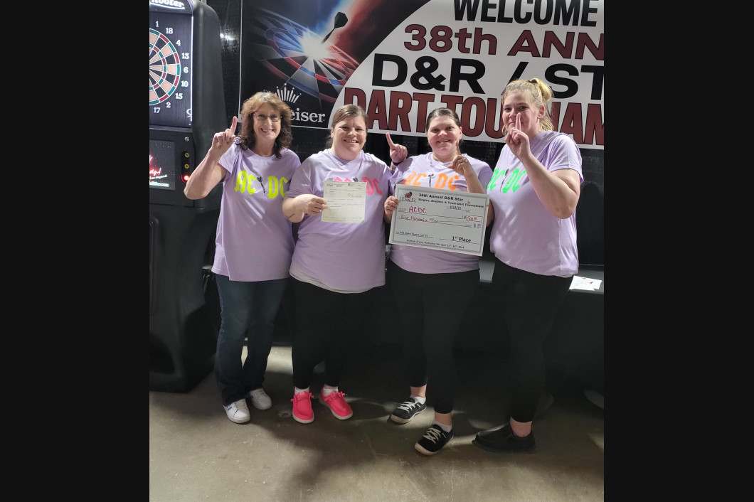 Four women in purple shirts hold certificates, celebrate a dart tournament, inside.