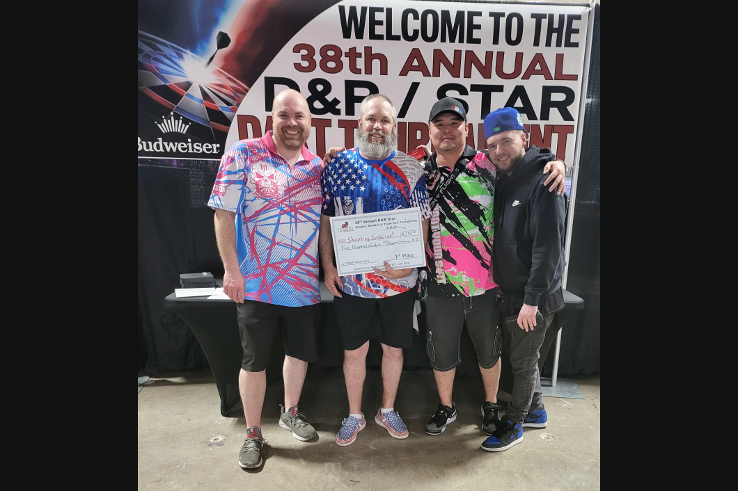 Four men holding certificate, posing at a darts tournament with colorful shirts, against a banner.