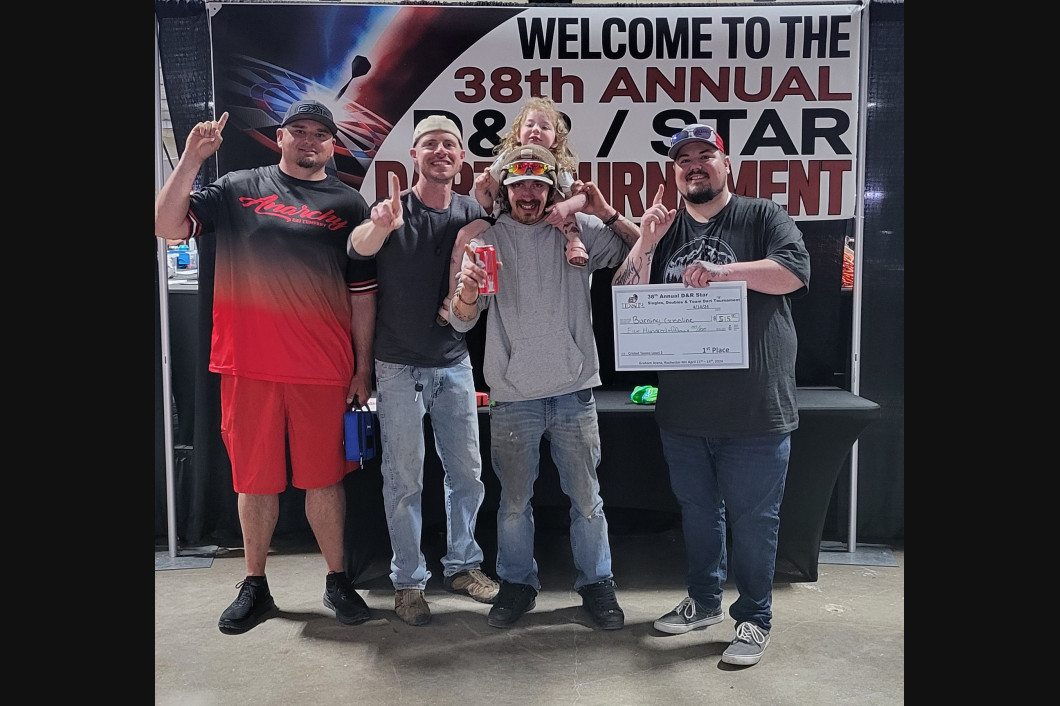 Four men celebrate winning a tournament; holding a check and prizes, posing with thumbs up. Event banner in background.