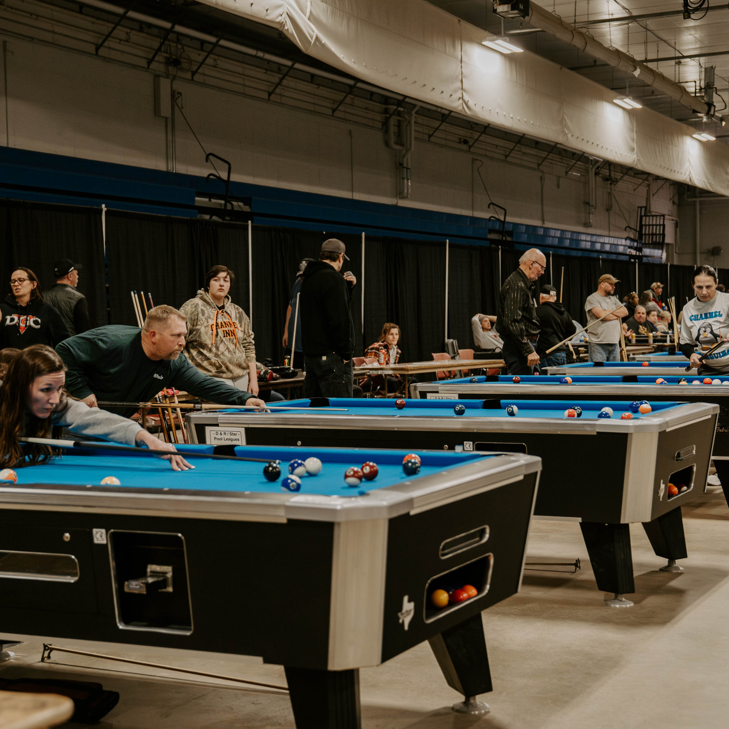 People playing pool at tables in a large indoor venue. Players line up shots, others watch. Blue felt tables.