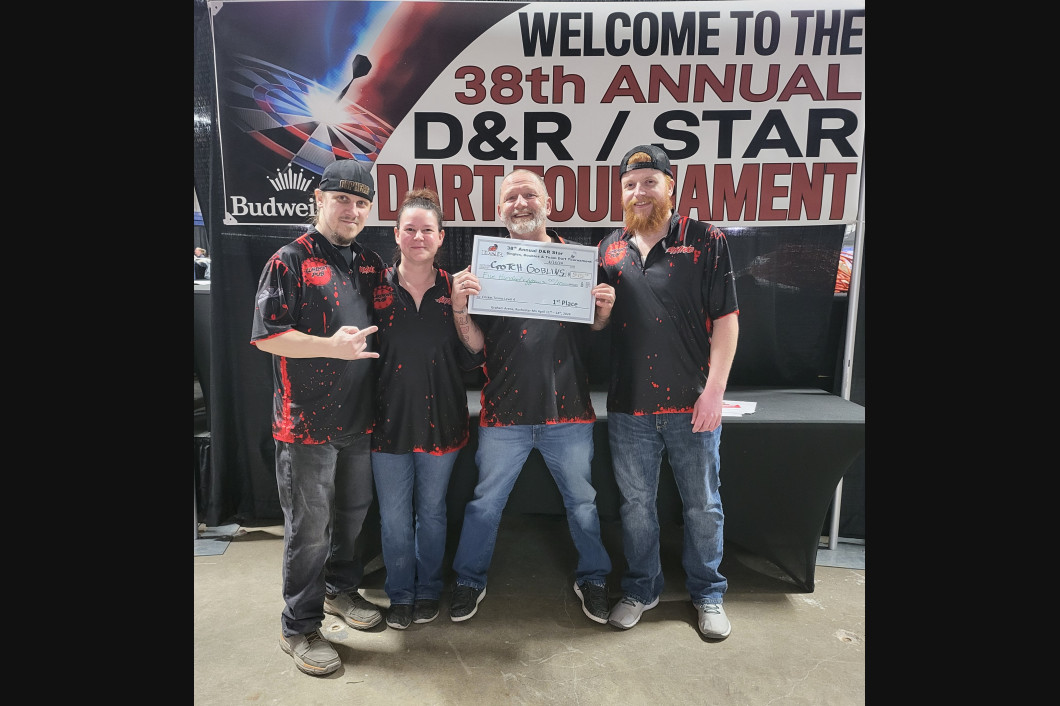 Four people holding a certificate, posing at the D&R/STAR Dart Tournament. They wear matching shirts. A banner is visible.