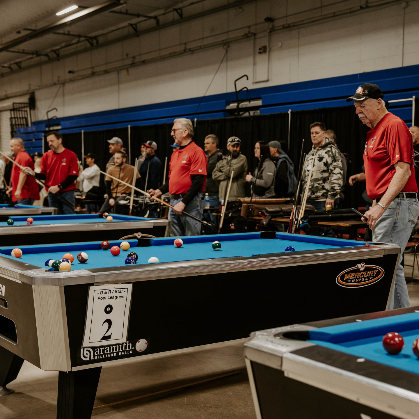 Pool tournament. People in red shirts line up to play at multiple pool tables indoors.