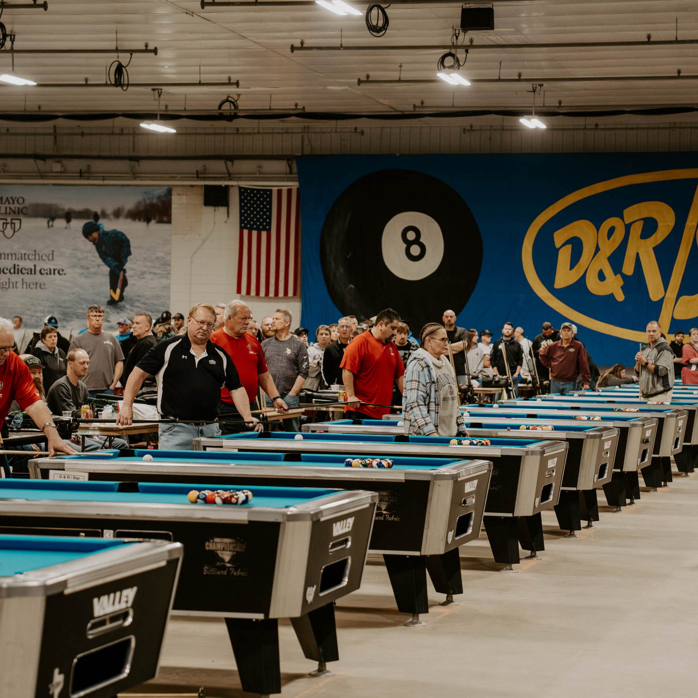 Pool tournament in a large hall with many players at blue tables. American flag, eight ball graphic.