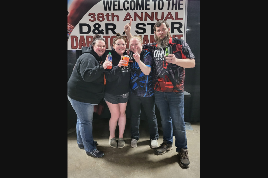 Four people hold trophies, celebrating at a darts tournament. 