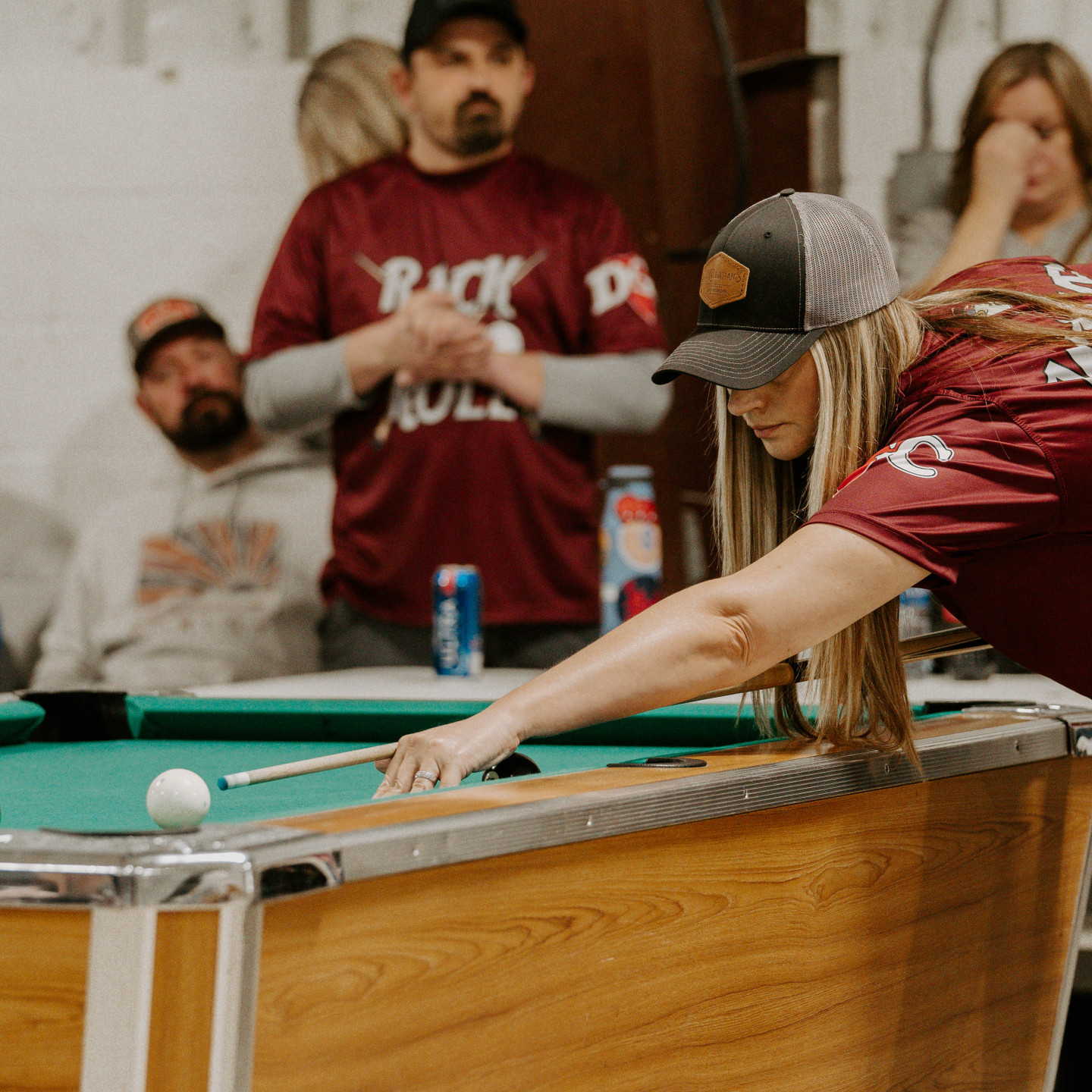 Woman in hat playing pool, aiming with cue stick. Spectators watch in the background.