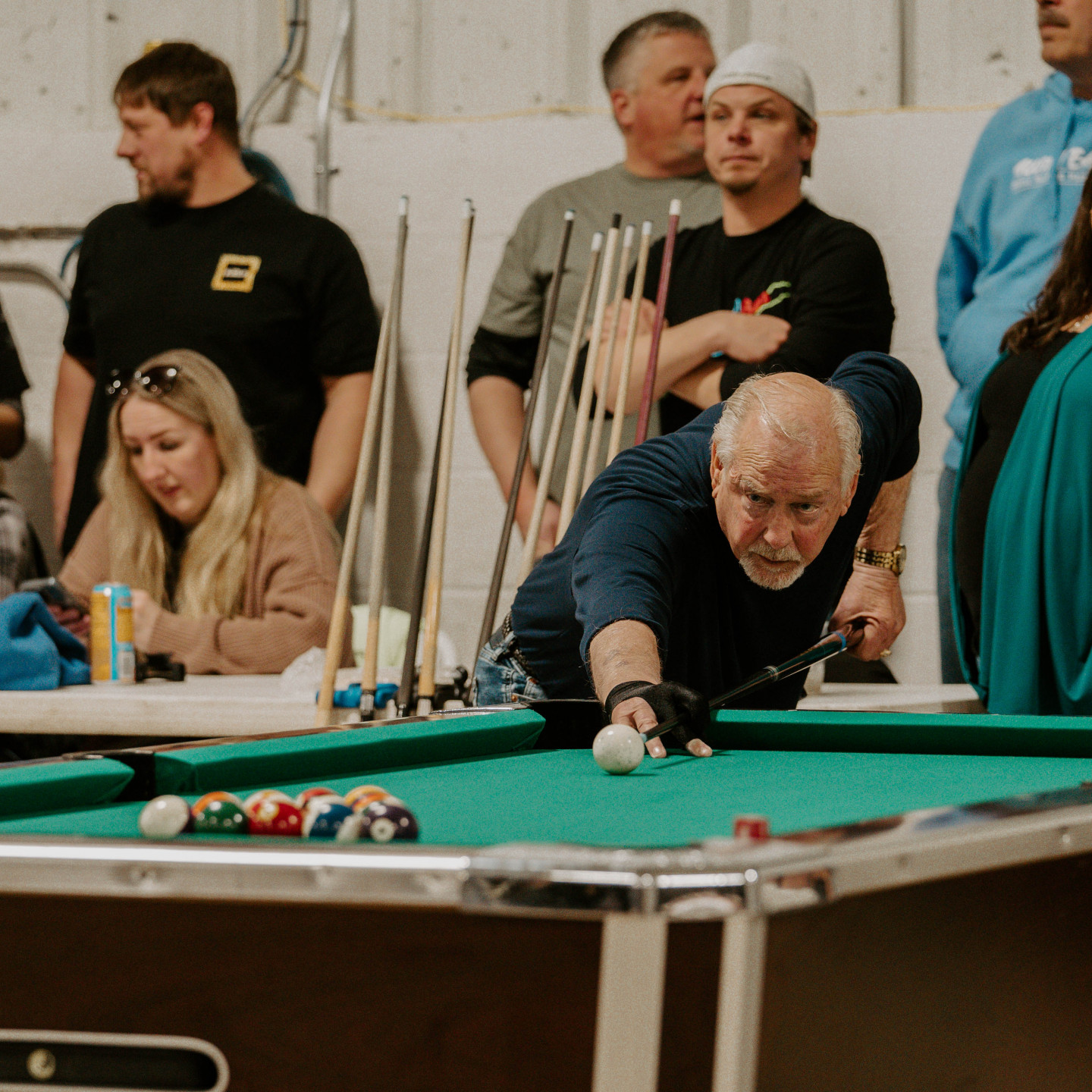 Man playing pool, aiming with cue. Others watch, some with cues. Green table, white walls.
