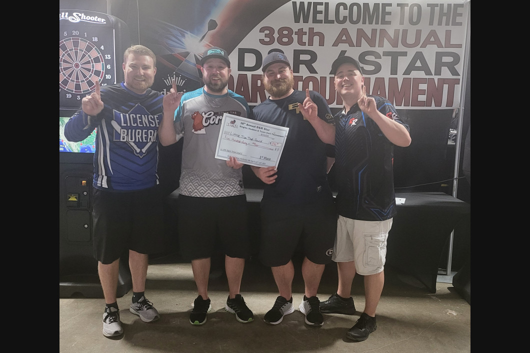Four men hold up fingers, posing with a large check, at a darts tournament. 