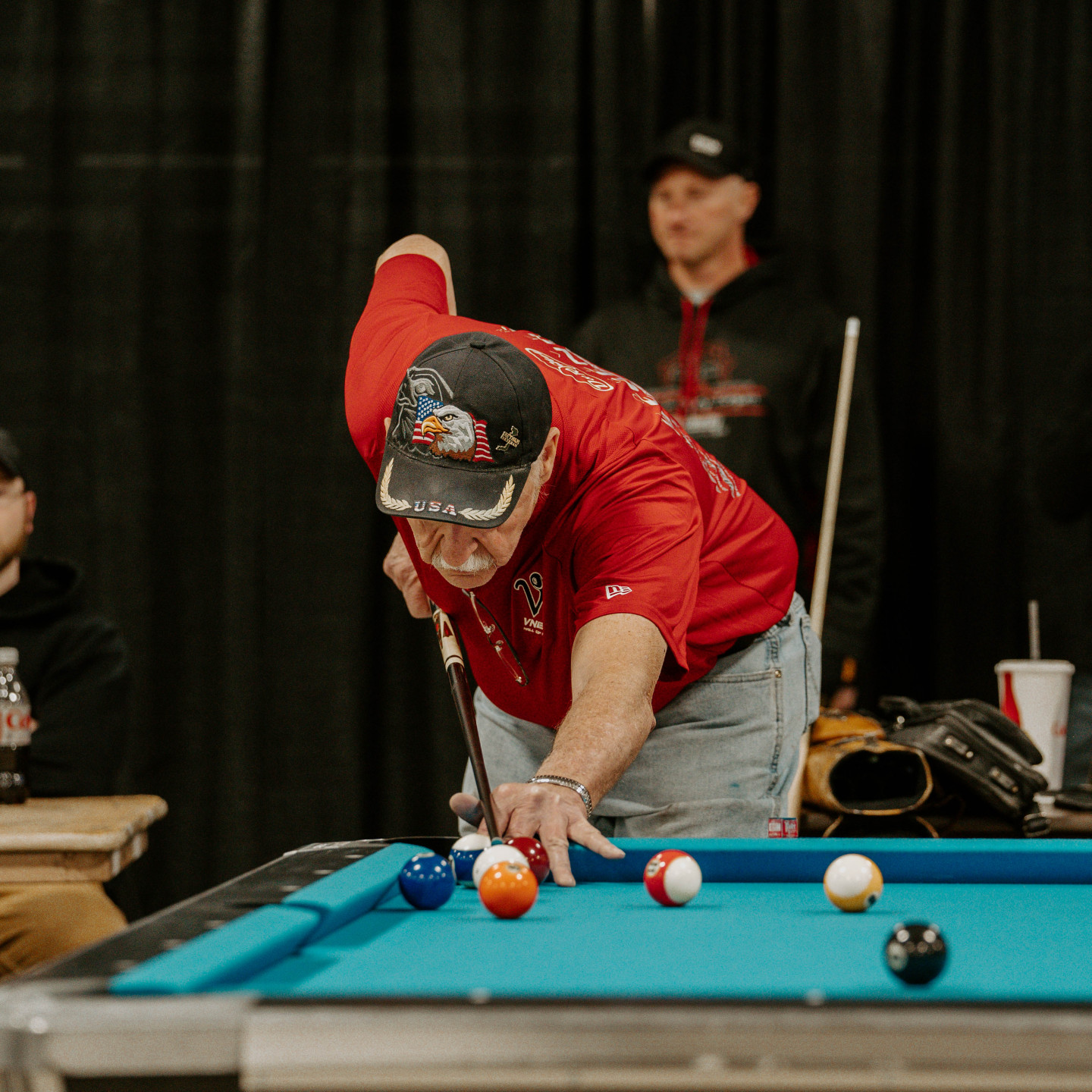 Man in red shirt playing pool, aiming with cue stick. Other man watches in the background.