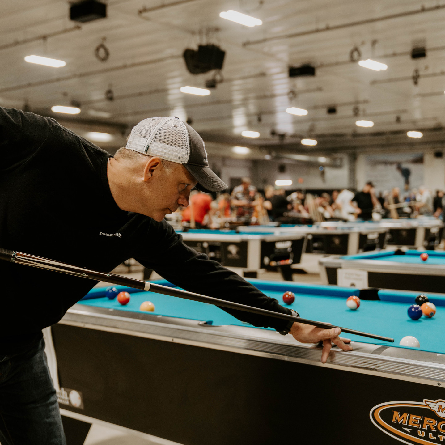 Man in hat lines up a pool shot at a tournament. Tables and spectators in background.