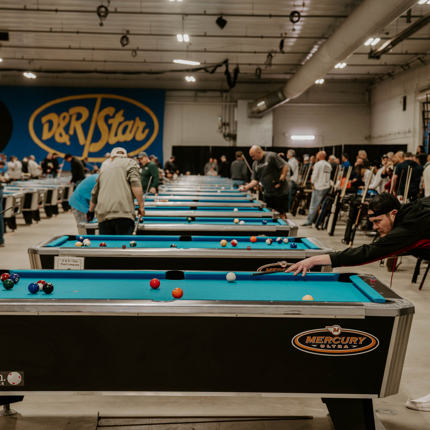 Pool tournament in large hall. Players line up at tables, shooting. 