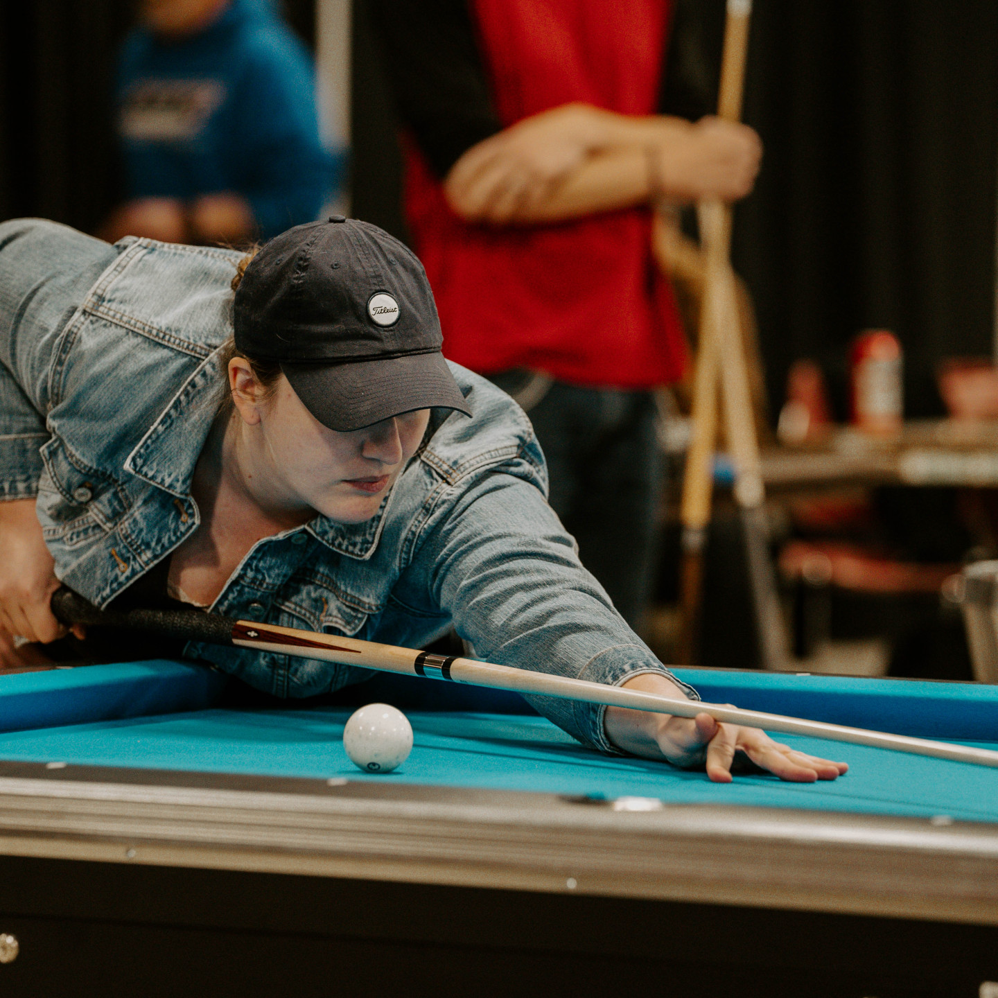 Woman in a denim jacket playing pool, aiming at the cue ball. Focus and determination on her face.