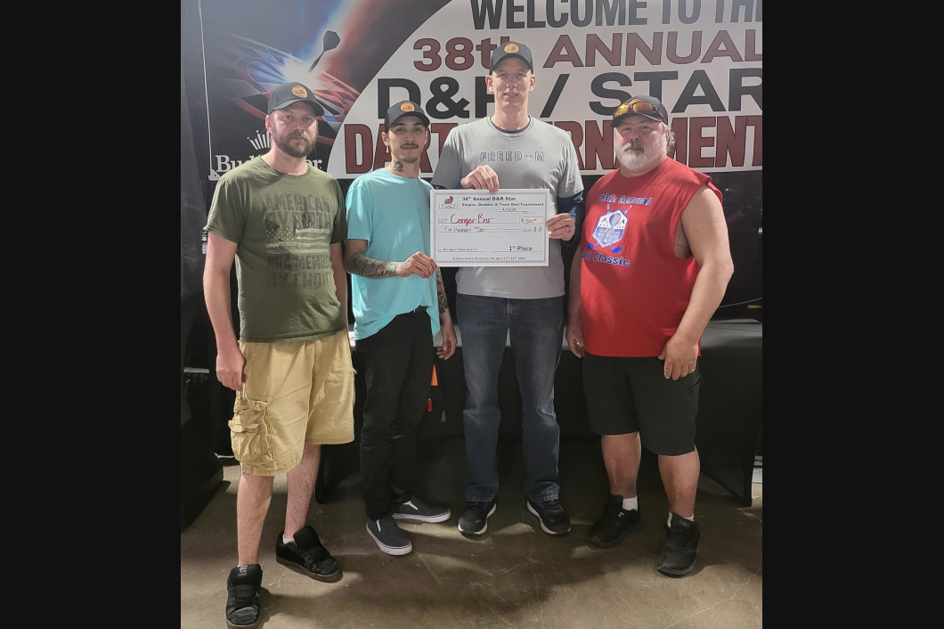 Four men holding a large check at a darts tournament; event banner in background.