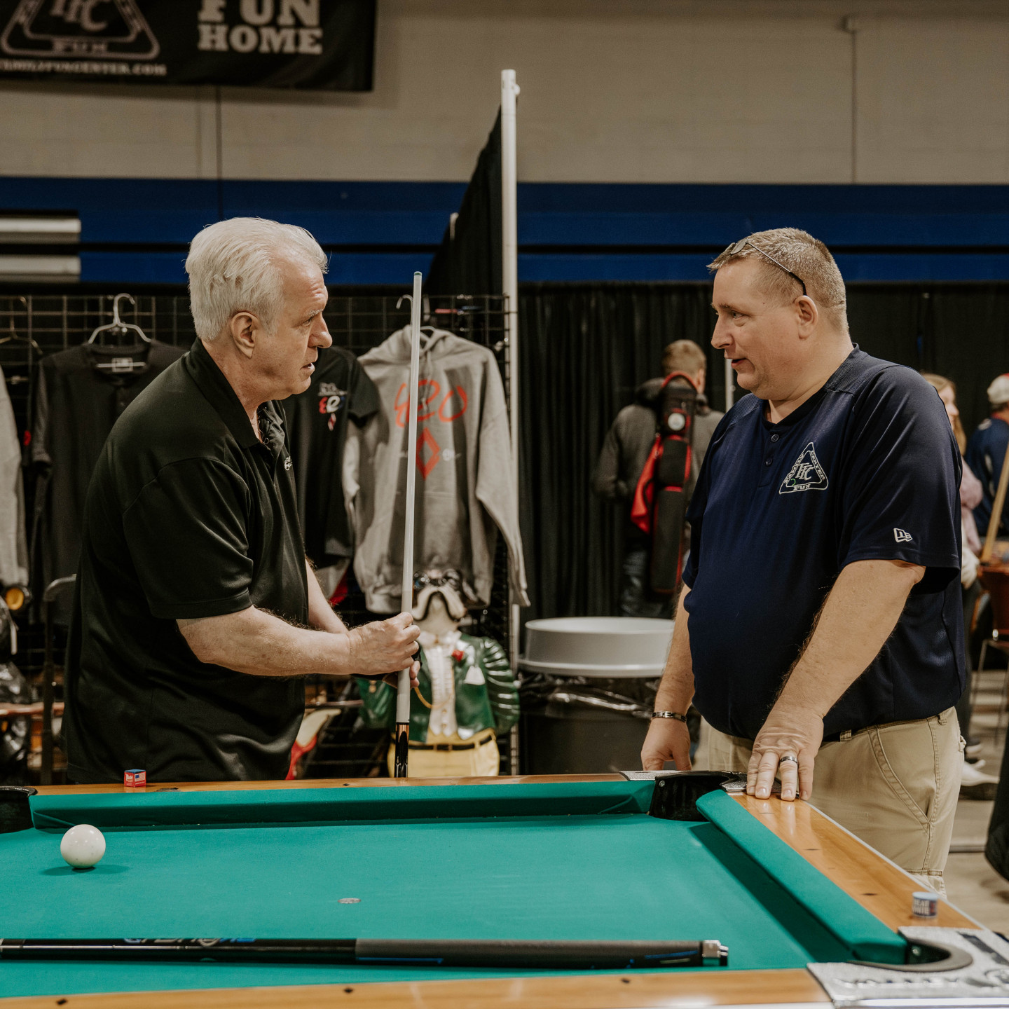 Two men converse beside a pool table. One points to something as the other listens. Indoor setting.