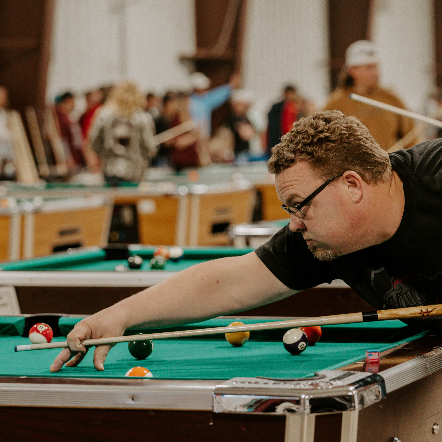 Man in black shirt lines up a pool shot, aiming cue at the ball on a green felt table.
