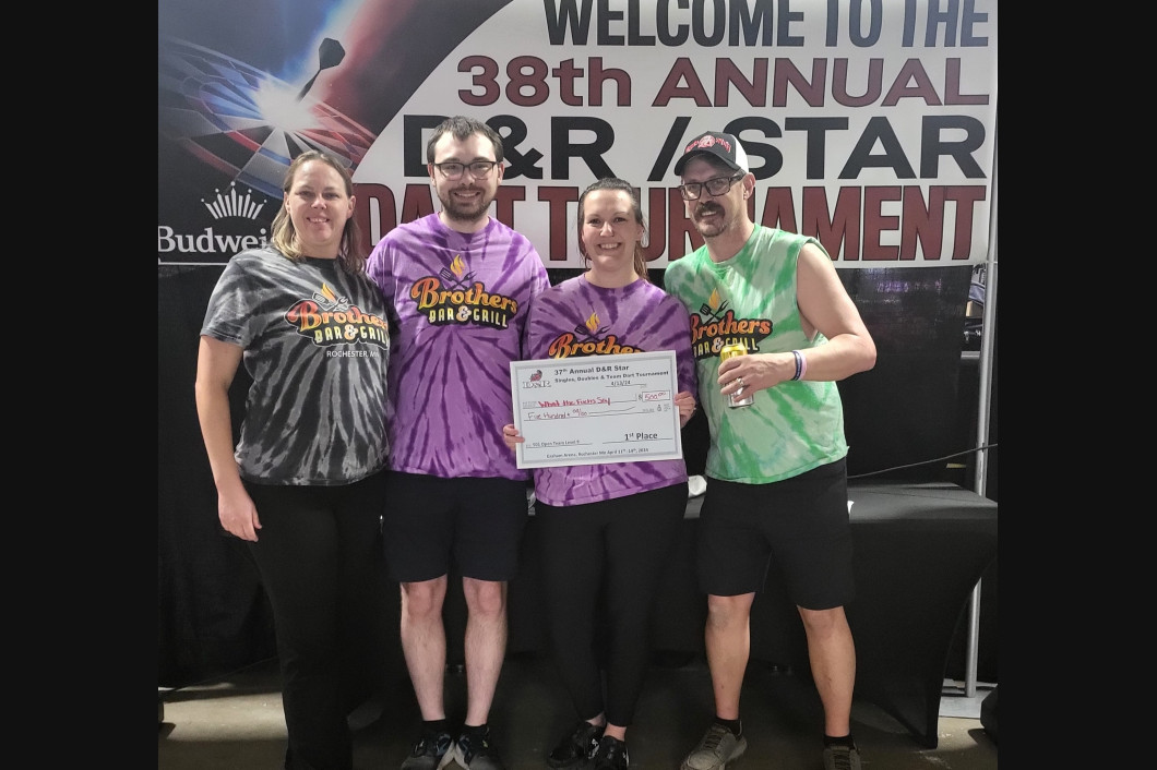 Four people holding a check at a tournament. Purple and green tie-dye shirts, event banner in background.