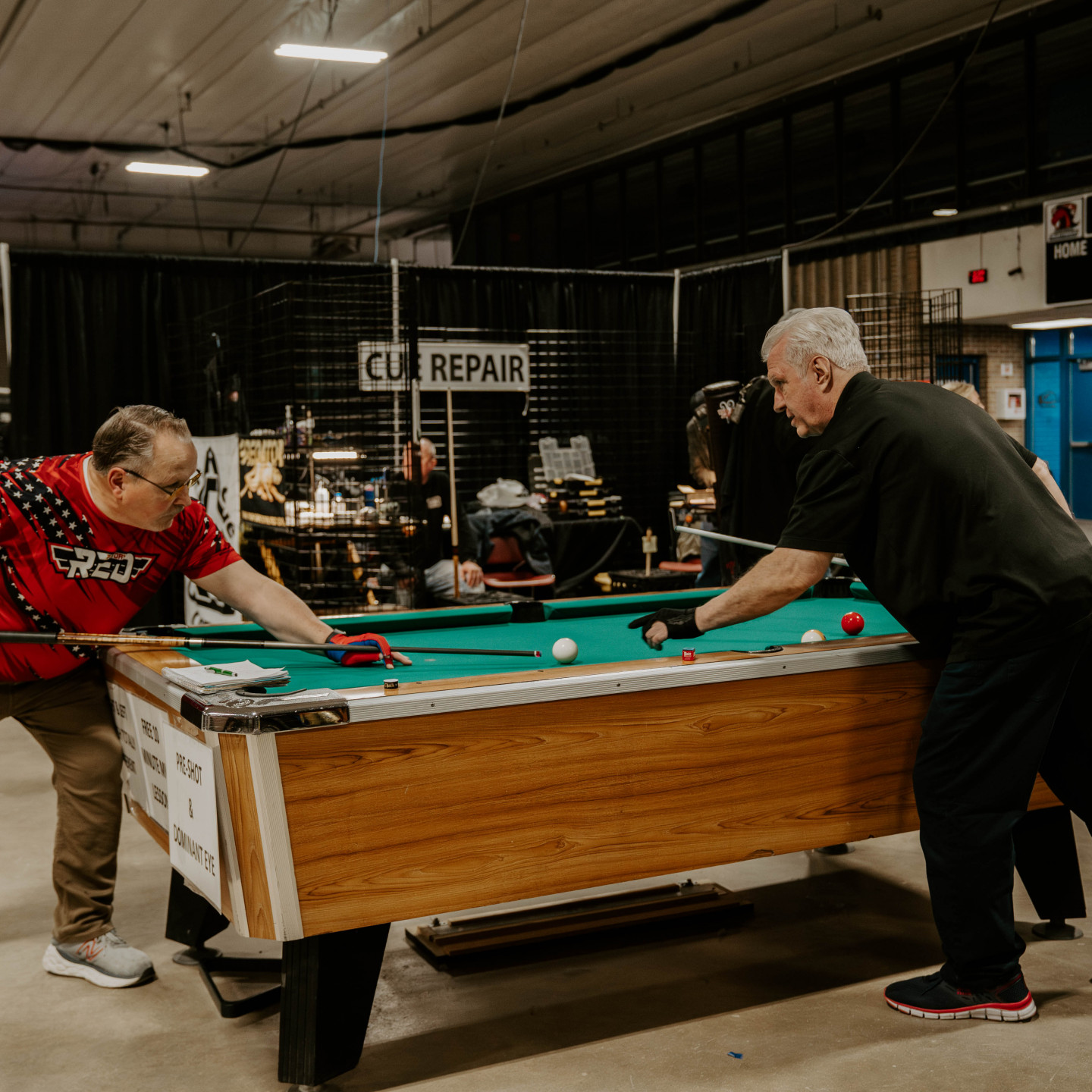 Two men playing pool at a cue repair shop. One wears a red shirt.