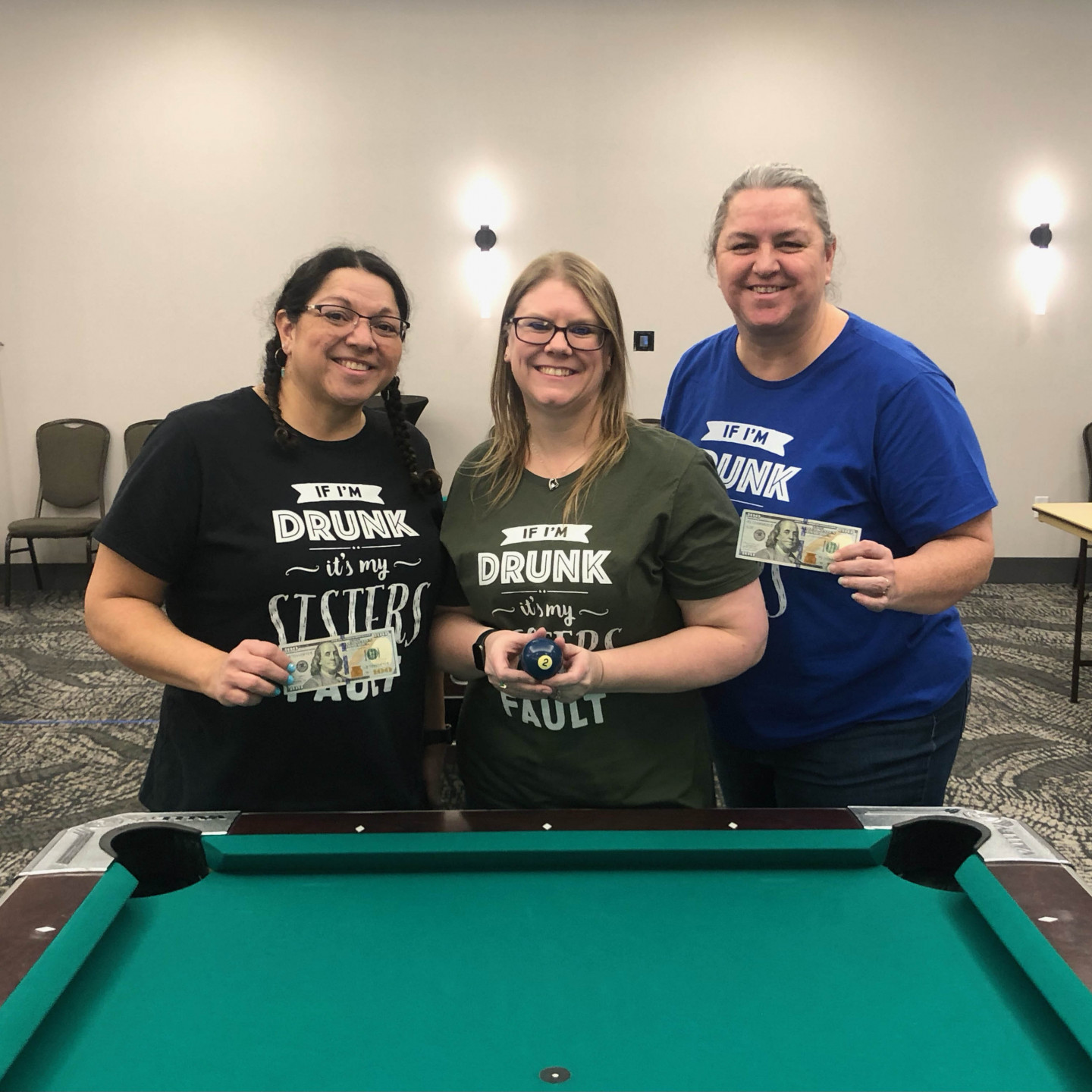 Three women holding cash and a billiard ball stand around a pool table. Each wears a t-shirt with a 