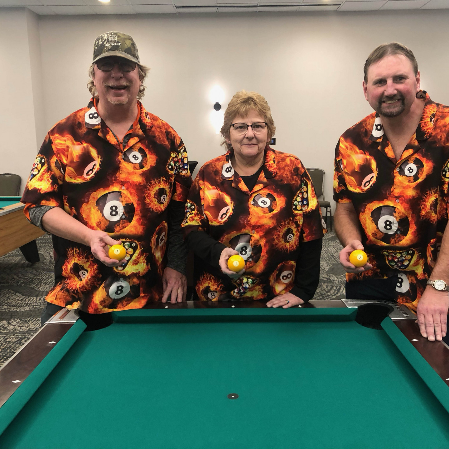 Three people in matching 8-ball shirts pose behind a pool table, each holding a yellow cue ball.