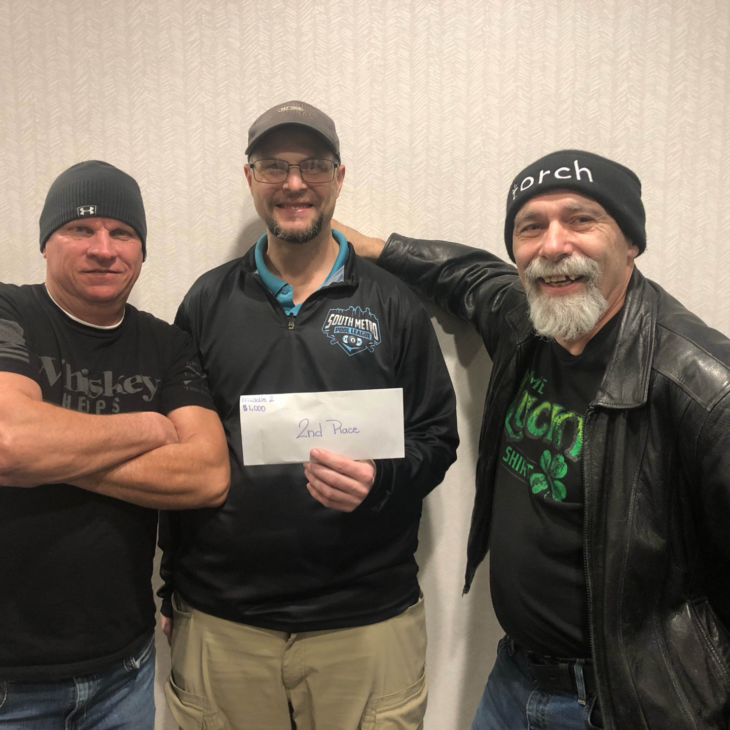 Three men stand, one holding a check.  Two wear hats. They pose in front of a neutral wall.