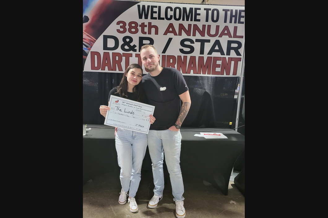 Two people pose with a large check at the 38th Annual D&R/Star Dart Tournament. They are standing near a table with a banner.