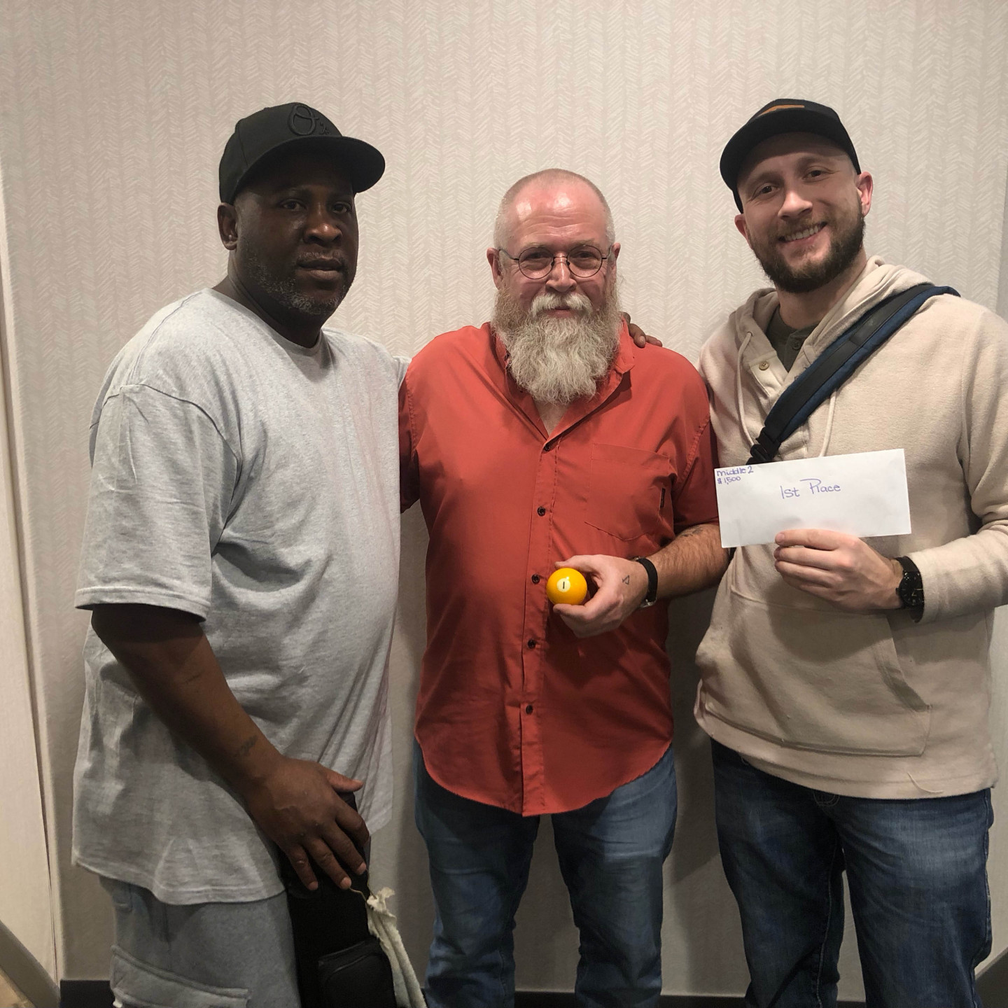 Three men pose for a photo. The central man holds a ball, the right man holds a check.