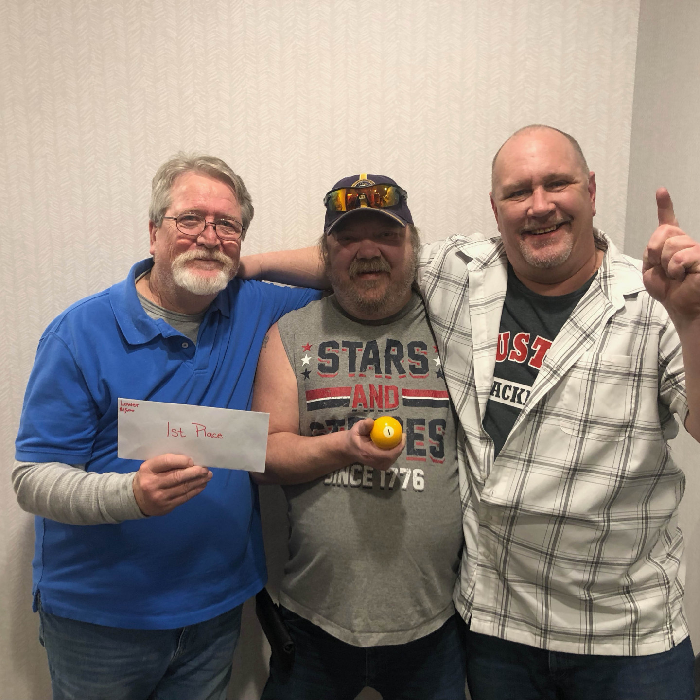 Three men pose, holding an envelope, a small fruit, and a raised finger. They stand in front of a white wall.