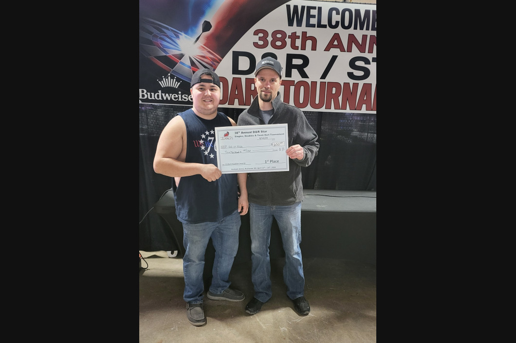 Two men holding a check at a tournament. One in a tank top, the other in a jacket, stand in front of a banner.