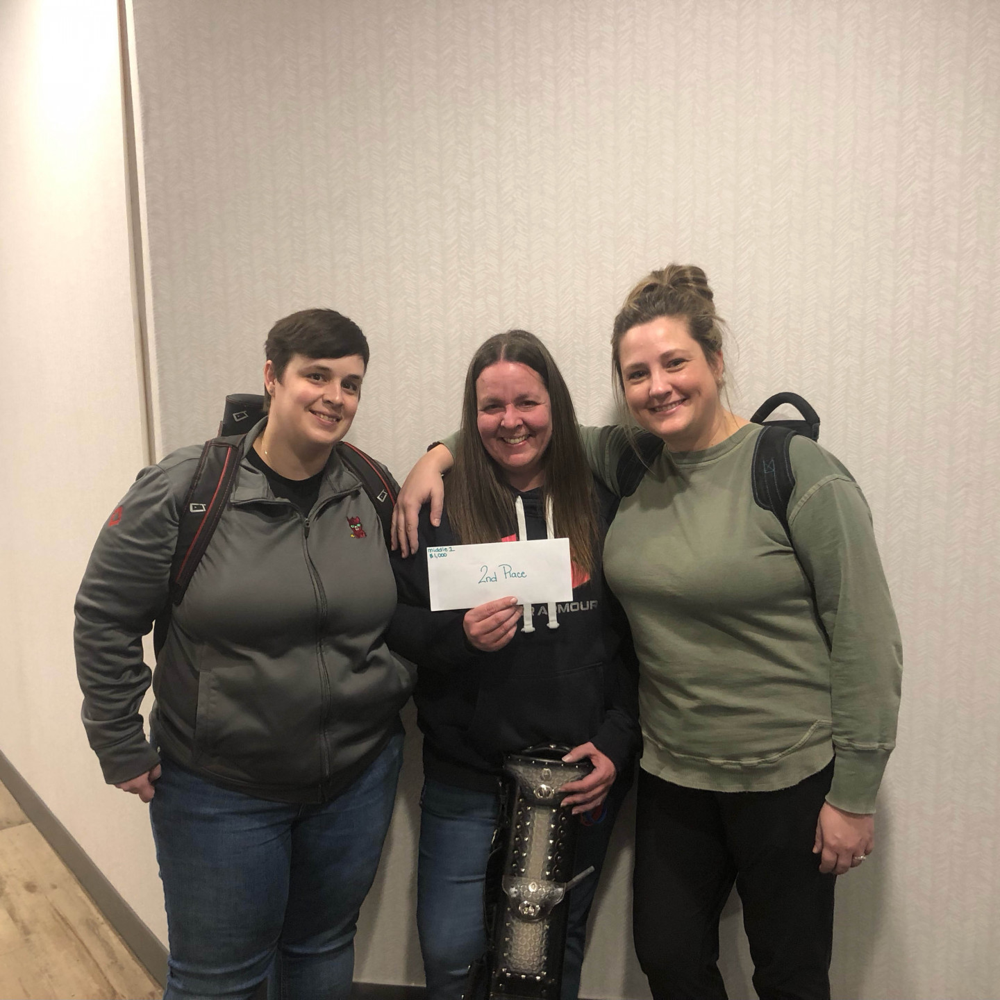 Three people stand side-by-side, one holding a check. They smile. Gray wall background.