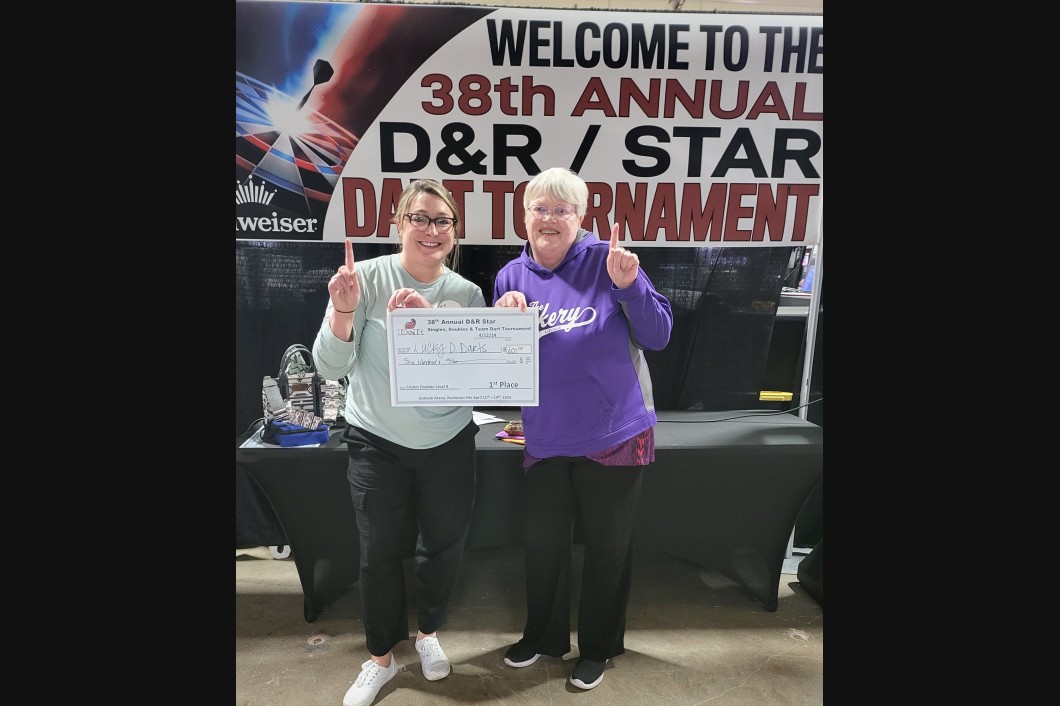 Two women at a darts tournament holding a check, thumbs up. The sign reads 