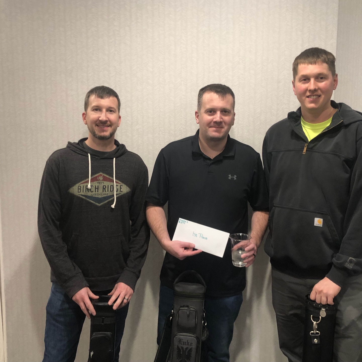 Three men stand indoors, one holding a check and a drink. The others hold what appears to be golf club bags.