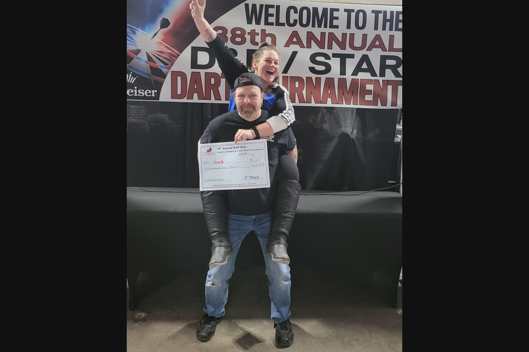 Man carrying a person, who is holding a certificate and celebrating, in front of a tournament banner.