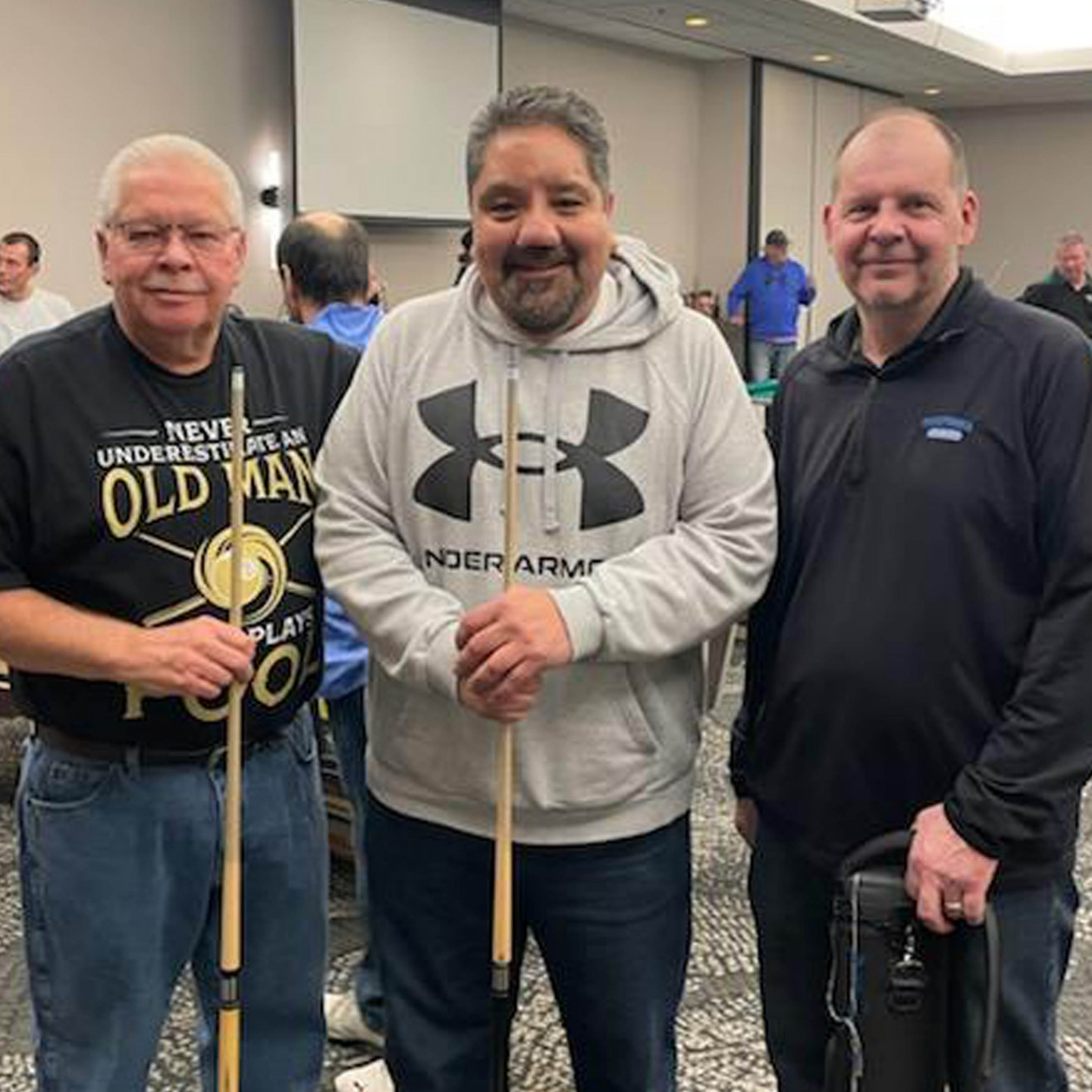 Three men with pool cues, smiling indoors. Two men wear casual hoodies. The other has an 