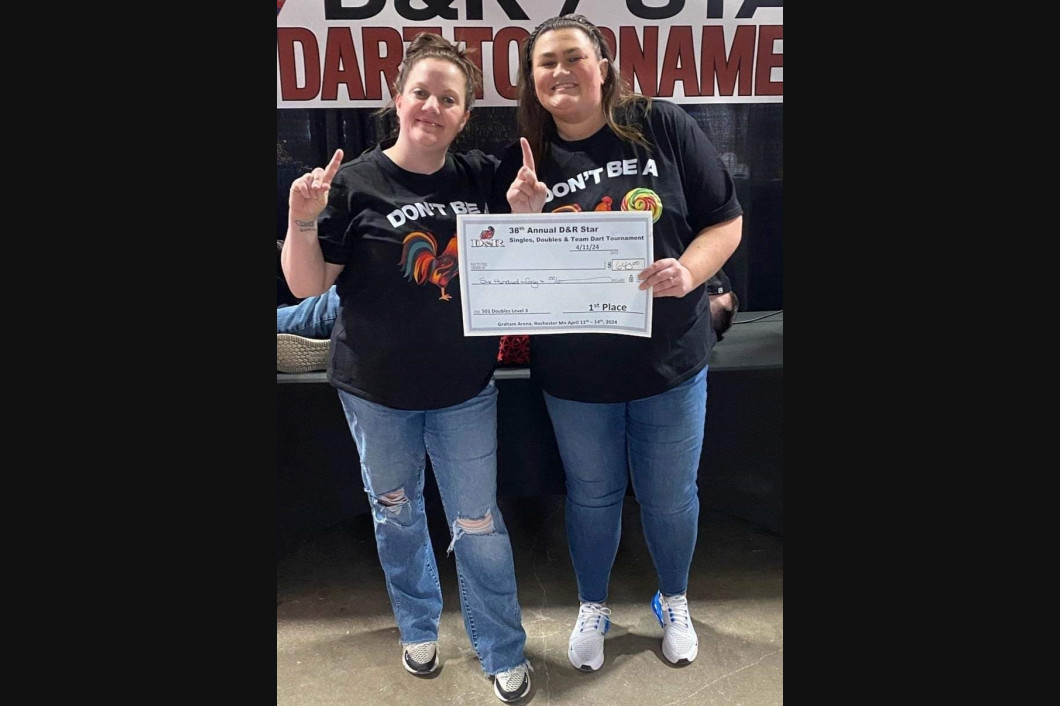 Two women hold a large check at a D & R dart tournament, both in black shirts and jeans.