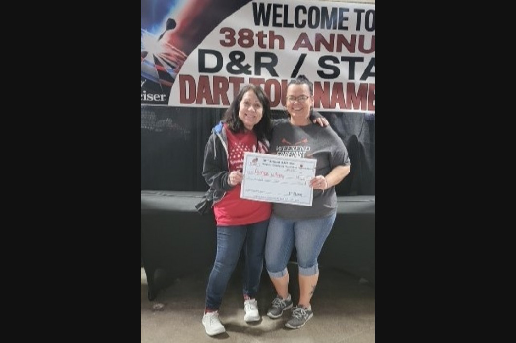 Two people hold a sign at a dart tournament. The banner says 