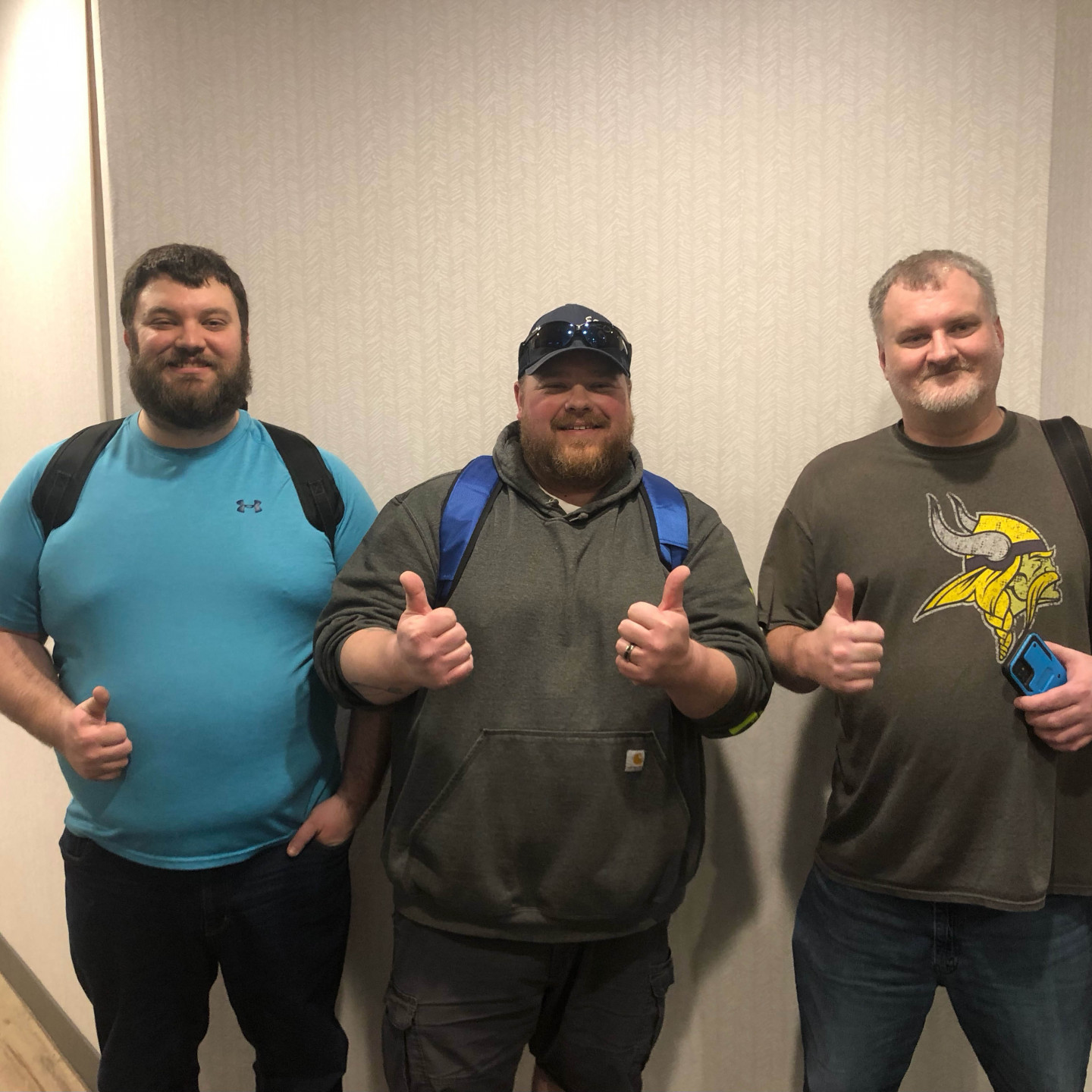 Three men giving thumbs up, standing in front of a light wall. They are wearing casual clothing and backpacks.