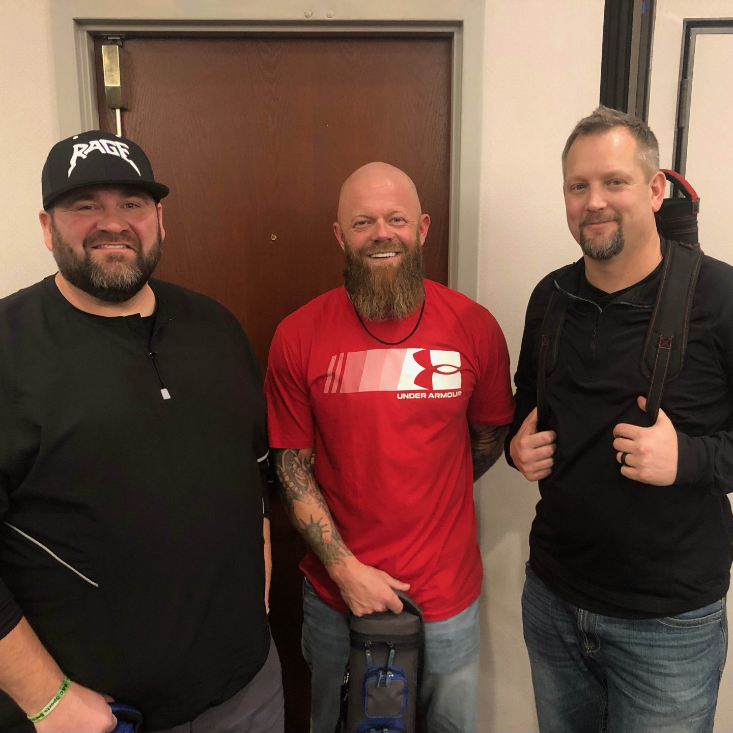 Three men smiling in front of a door. The middle man holds a guitar case, while the man on the right carries a guitar on his back.