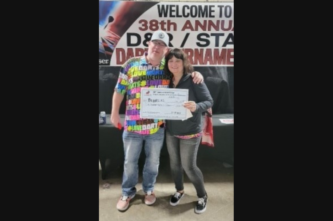 Man and woman holding a check at a dart tournament. They are posing in front of a banner.