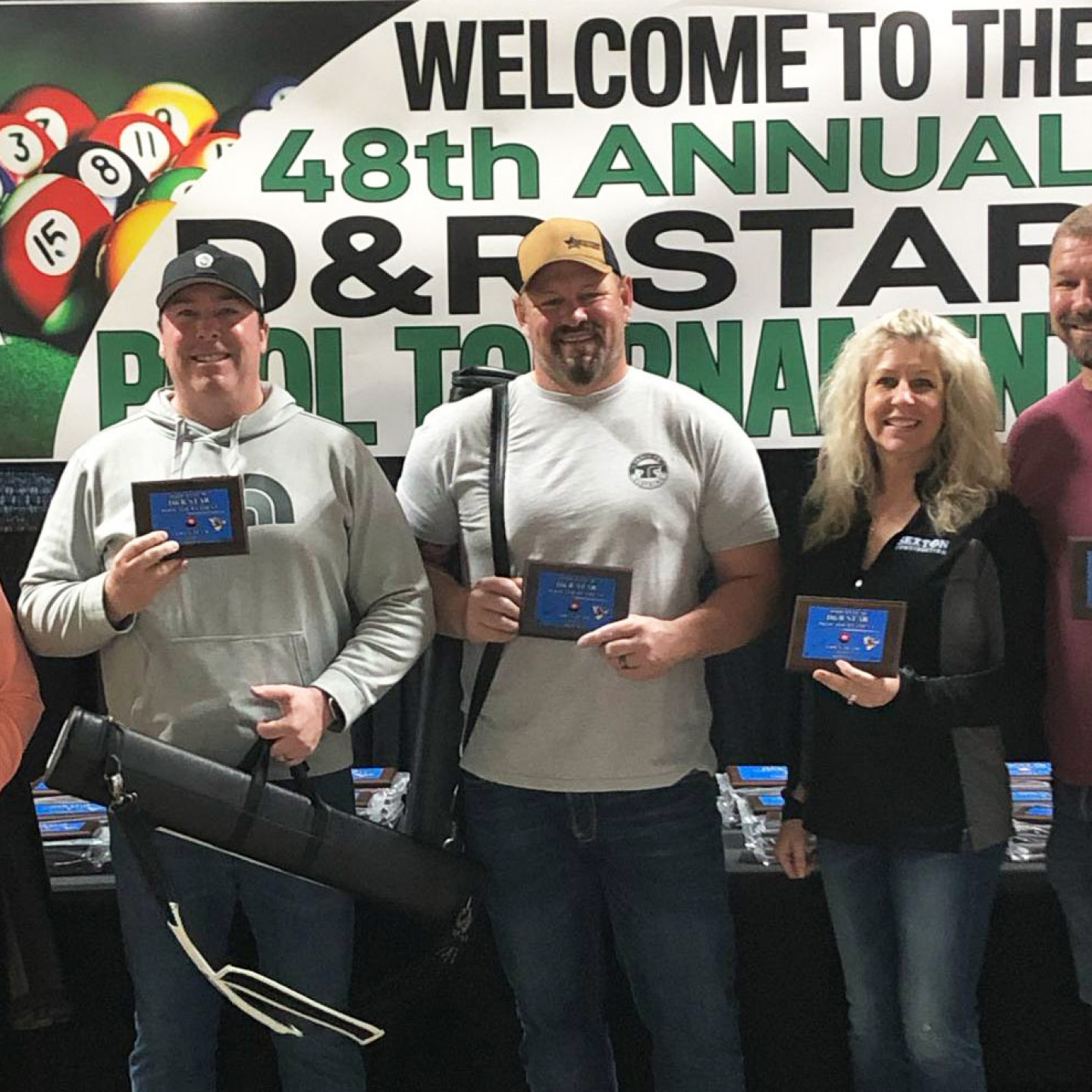 Pool tournament winners holding trophies, standing in front of a banner that reads 