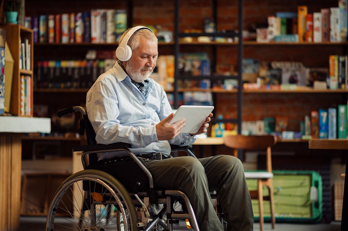 An elderly man in a wheelchair is wearing headphones and using a tablet.
