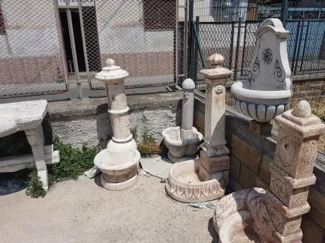 Vari ornamenti da giardino in pietra e elementi per fontane esposti su un'area esterna sabbiosa vicino al muro di un edificio.