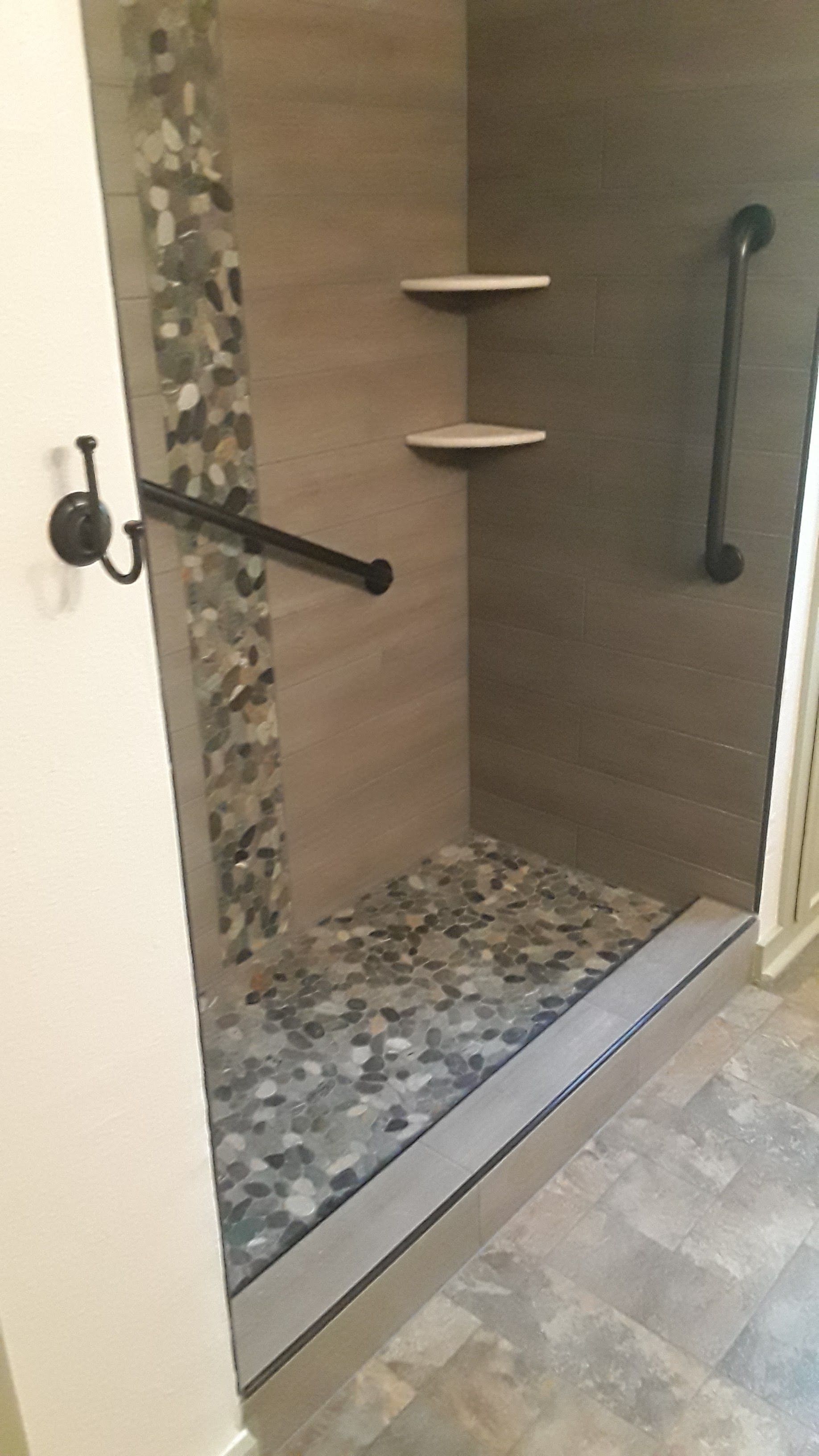 Walk-in shower with pebble tile floor, wood-look walls, corner shelves, and black grab bars
