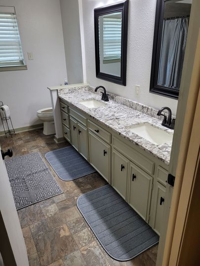Bathroom vanity with double sinks, mirrors, and gray rugs on a patterned tile floor.