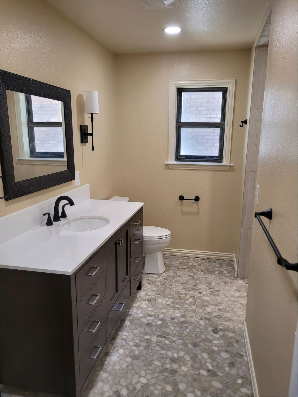 Narrow beige bathroom with dark vanity, white sink, wall mirror, toilet, and window at the end.