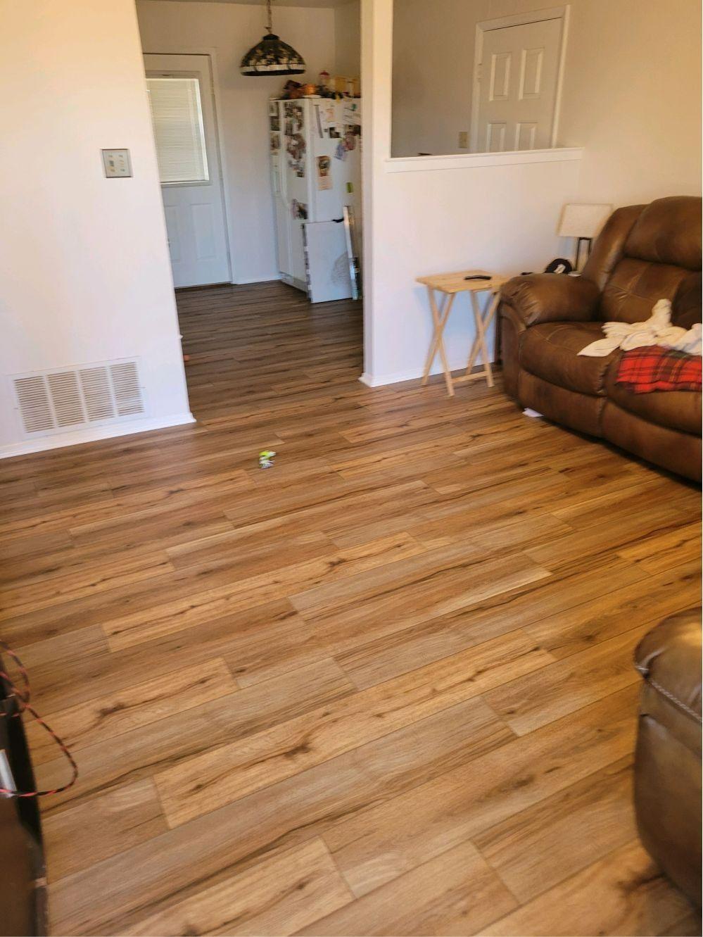 Empty living room with wood floors, brown recliner, and a doorway to a kitchen in the background