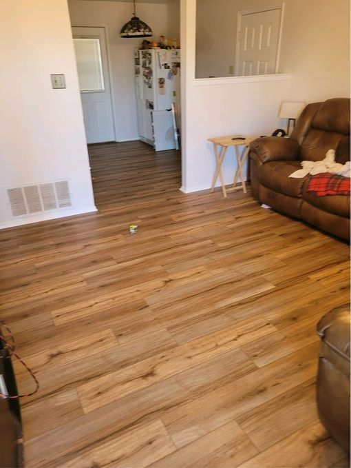 Empty living room with wood floors, brown recliner, and a doorway to a kitchen in the background