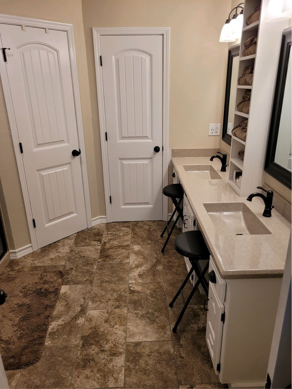 Narrow bathroom with two white doors, double sink vanity, black fixtures, and tan tile floor.