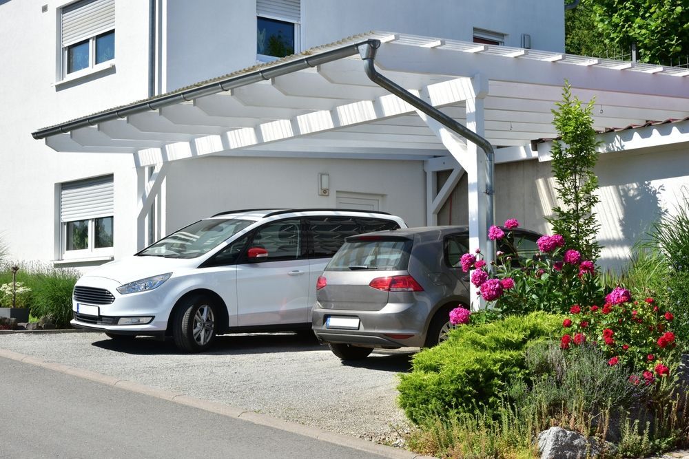 Two cars parked under a white carport attached to a white building with landscaping.
