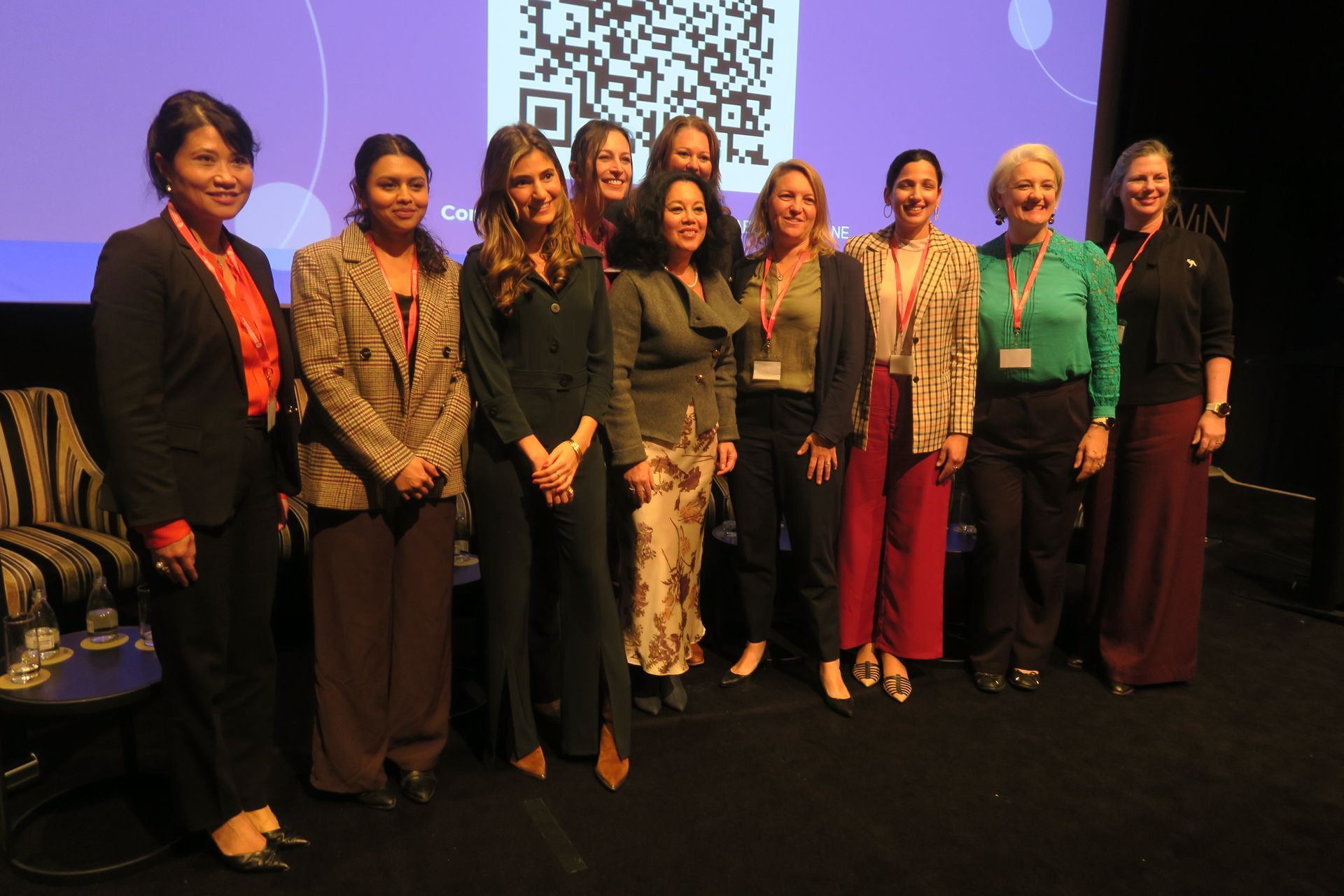 Group of women standing on stage, posing. Purple backdrop with QR code.